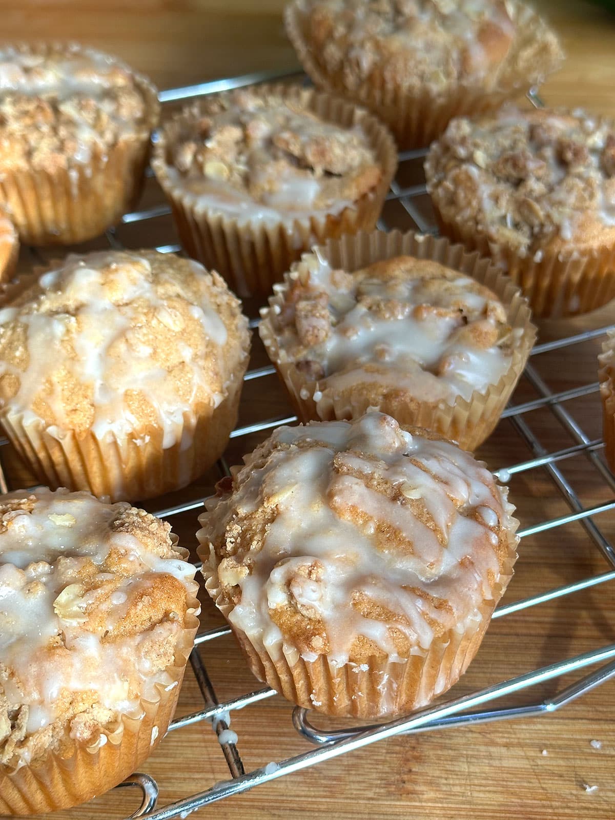 Vegan apple muffins with crumb topping and glaze sit on a metal cooling rack on a wooden surface, illuminated by natural light.