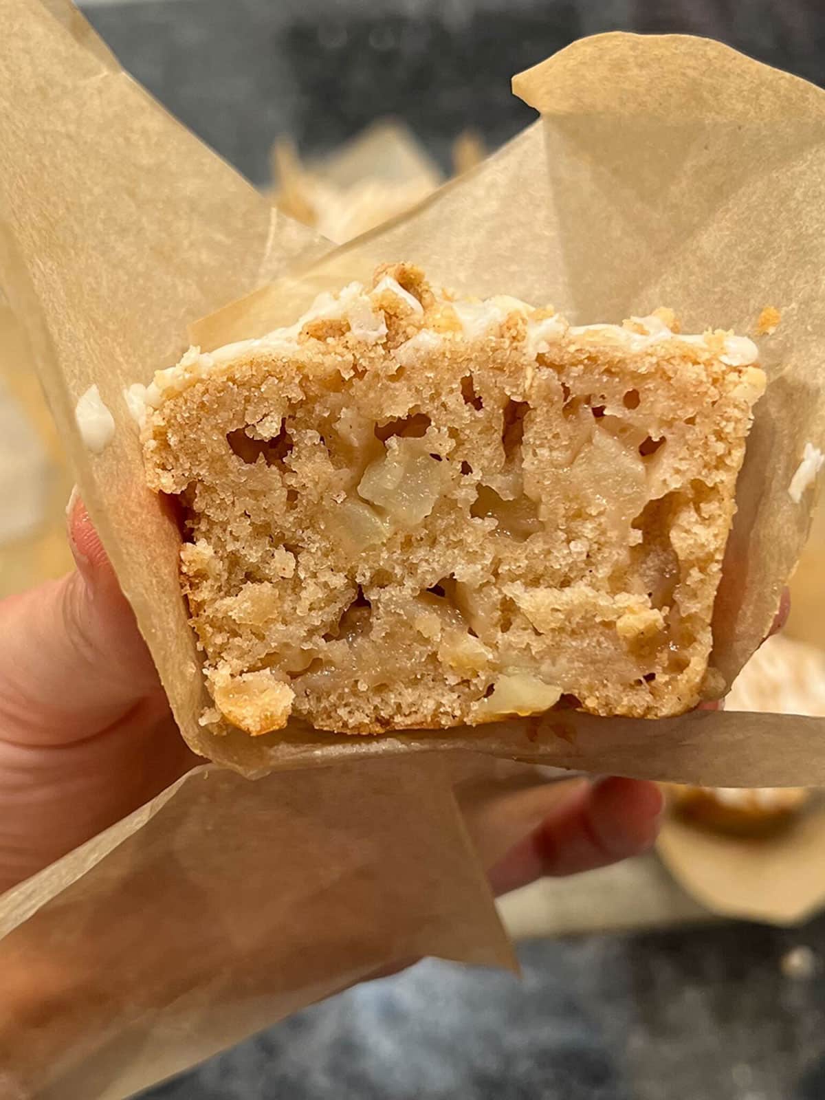 A hand holds a square piece of cake with visible apple chunks and a golden, moist crumb—reminiscent of vegan apple muffins—wrapped in brown parchment paper.