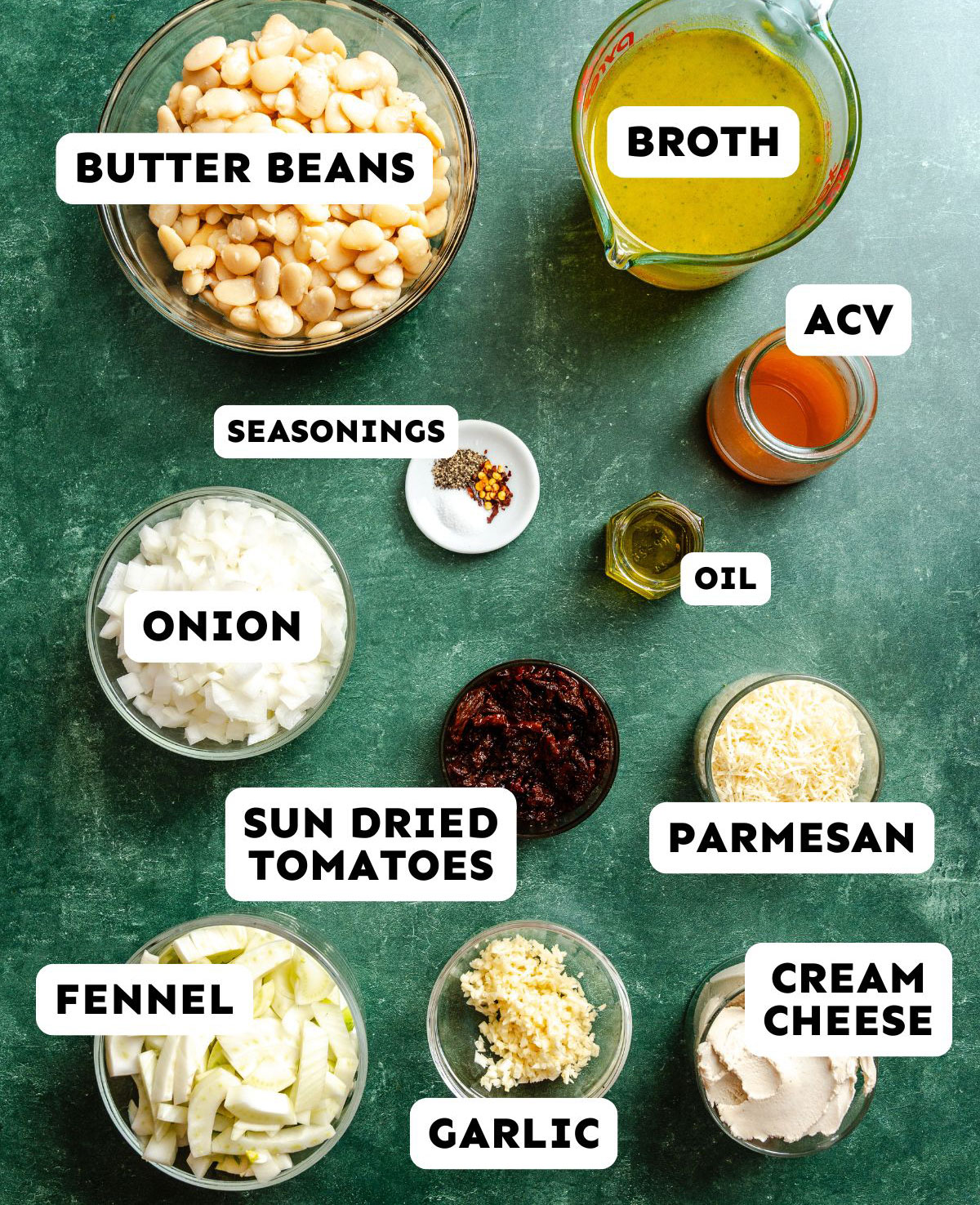 Ingredients for a dish are arranged on a green surface, including butter beans, broth, apple cider vinegar, oil, onion, seasonings, sun dried tomatoes, parmesan, fennel, garlic, and cream cheese.