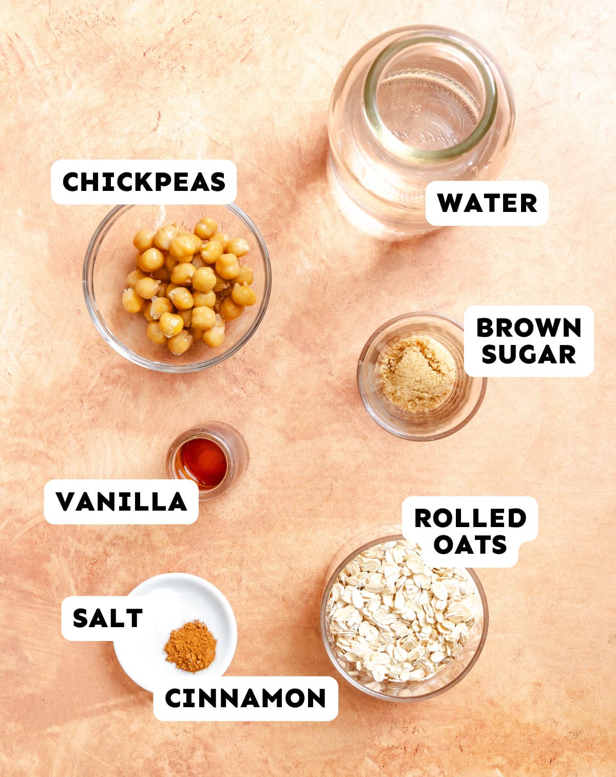 A flat lay of ingredients in bowls and jars: chickpeas, water, brown sugar, rolled oats, vanilla, salt, and cinnamon, each labeled with text.