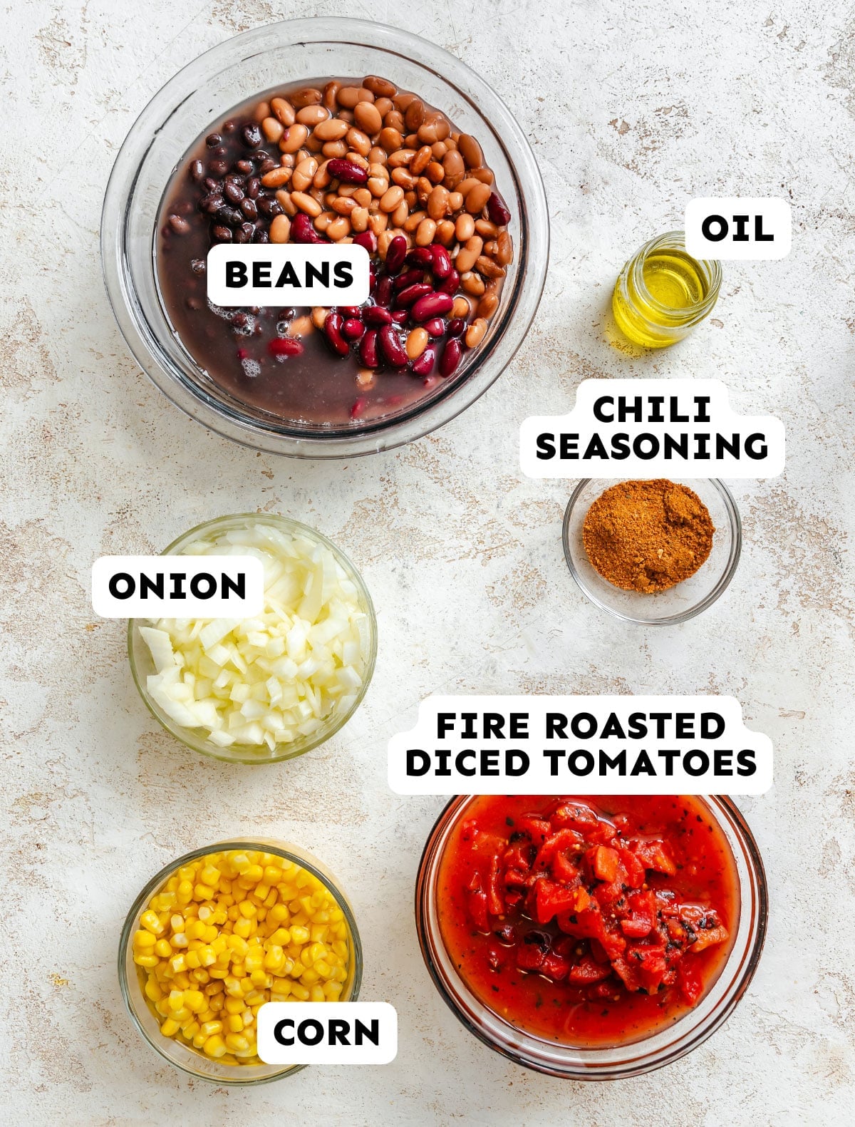 Five bowls containing beans, diced onion, corn, fire roasted diced tomatoes, chili seasoning, and a small container of oil arranged on a light surface, each ingredient labeled.