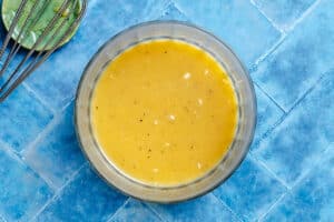 A glass bowl filled with yellow vinaigrette dressing sits on a blue tile surface next to a metal whisk and a small green dish.