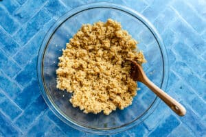 A clear glass bowl filled with cooked quinoa sits on a blue patterned surface, with a wooden spoon resting inside the bowl.