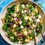 A bowl of quinoa salad with chopped kale, red onions, raisins, and feta cheese, served with two forks.