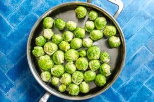 Brussels sprouts in a stainless steel pan filled with water, placed on a bright blue tiled surface.