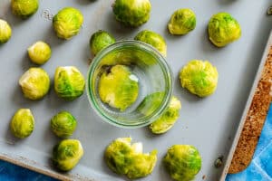 A baking sheet with whole Brussels sprouts, some of which are being flattened by a glass jar placed in the center.