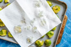 A baking sheet with flattened Brussels sprouts, partially covered by a sheet of paper towel, sits on a blue surface.