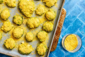 A baking sheet with smashed Brussels sprouts topped with yellow nutritional yeast, next to a small glass bowl of nutritional yeast, on a blue tiled surface.