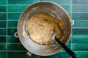 A metal mixing bowl containing thick, beige batter is placed on a green tiled countertop, with a black spoon resting inside the bowl.