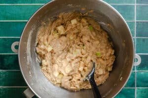 A metal mixing bowl with apple cake batter and a black spatula on a green tiled countertop.