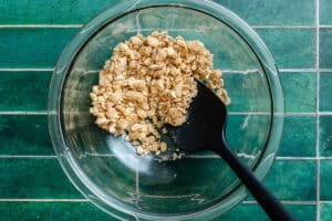 A glass bowl containing a mixture of oats and sugar, with a black spatula resting inside, placed on a green tiled surface.