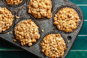 A muffin tin with six baked muffins topped with a crumbly oat streusel, resting on a green tiled surface.
