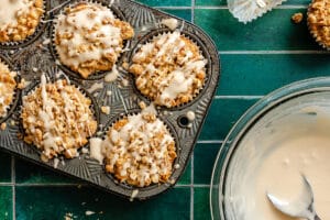 A metal muffin tin with freshly baked muffins drizzled with glaze sits on a green tiled surface beside a glass bowl with leftover glaze and a spoon.