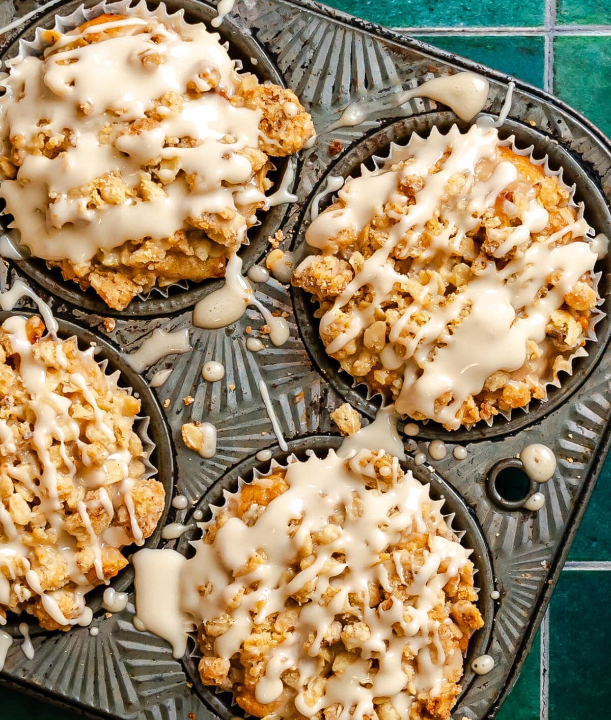 Four vegan apple muffins with crumb topping and white glaze are shown in a muffin tin on a green tiled surface.