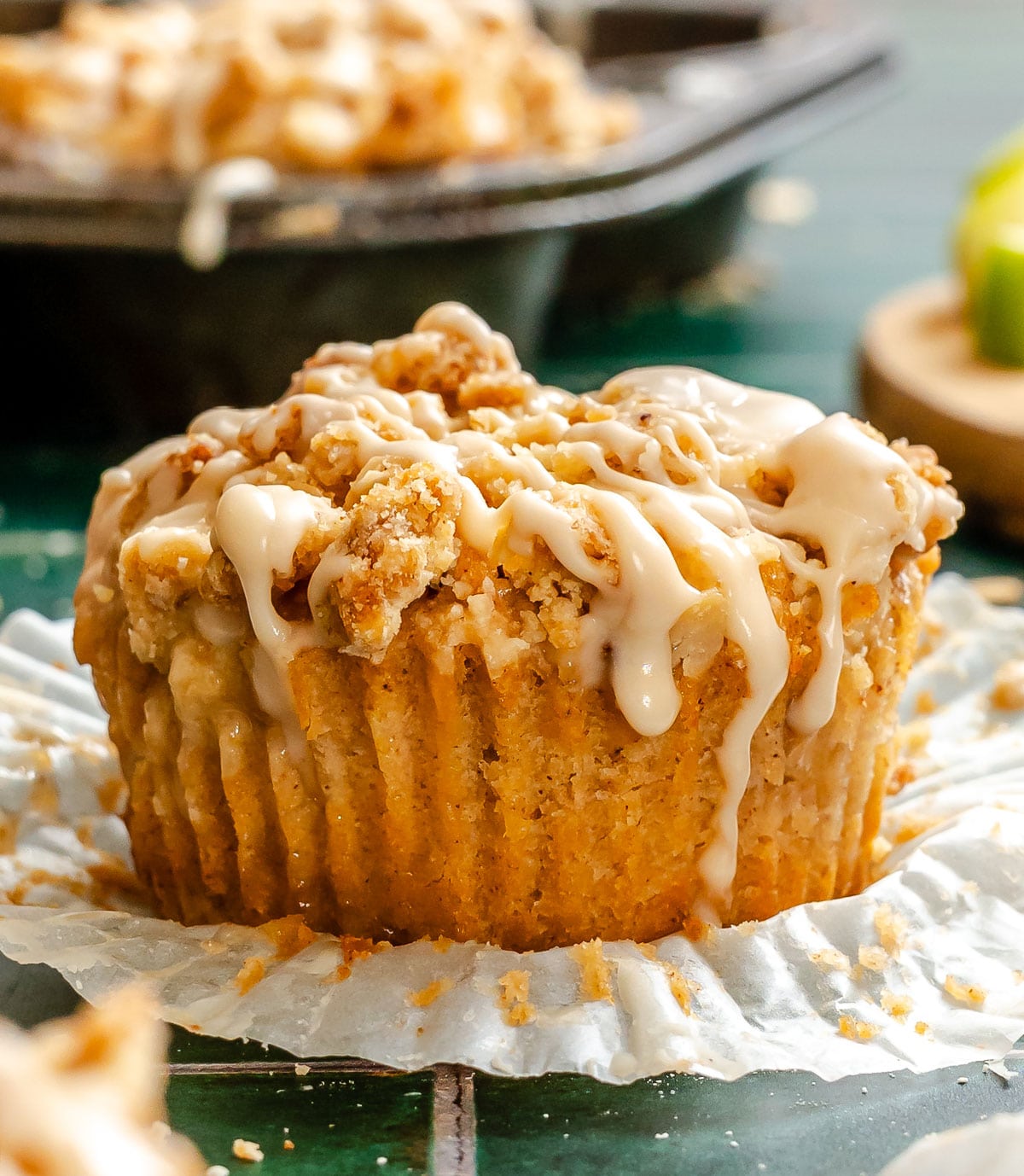 A close-up of a muffin with crumb topping and white icing drizzle, resting on a peeled-back paper liner. A muffin tin is blurred in the background.