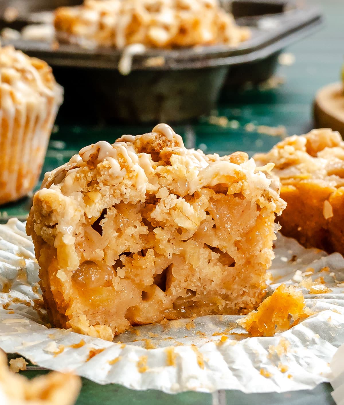 A close-up of a muffin with a crumb topping and white icing, sliced in half on a muffin wrapper, showing a moist apple filling inside. Other muffins are blurred in the background.