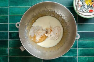 A metal mixing bowl containing sugar, milk, vanilla extract, and what appears to be cream cheese sits on a green tiled surface beside a whisk and small bowl.