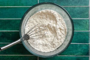 A glass bowl filled with flour mixture and a metal whisk, placed on a green tiled surface.