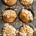 Six vegan apple muffins with crumble topping and white glaze, arranged in a metal muffin tin.