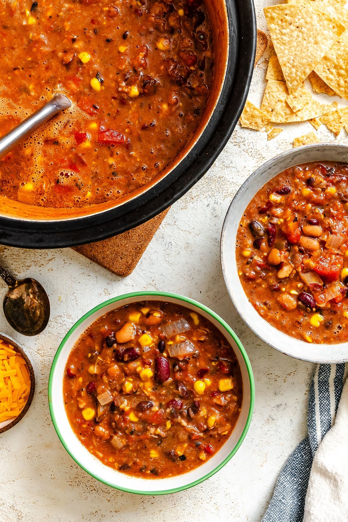Three bowls of vegetarian chili with beans, corn, and tomatoes, served with tortilla chips and shredded cheese on a light surface.