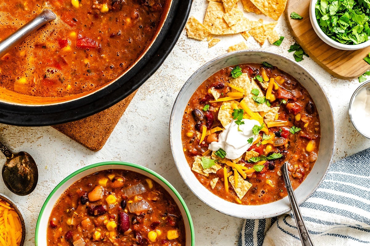 Overhead view of bowls of chili topped with sour cream, shredded cheese, tortilla chips, and cilantro, with a pot of chili and bowls of garnishes nearby.