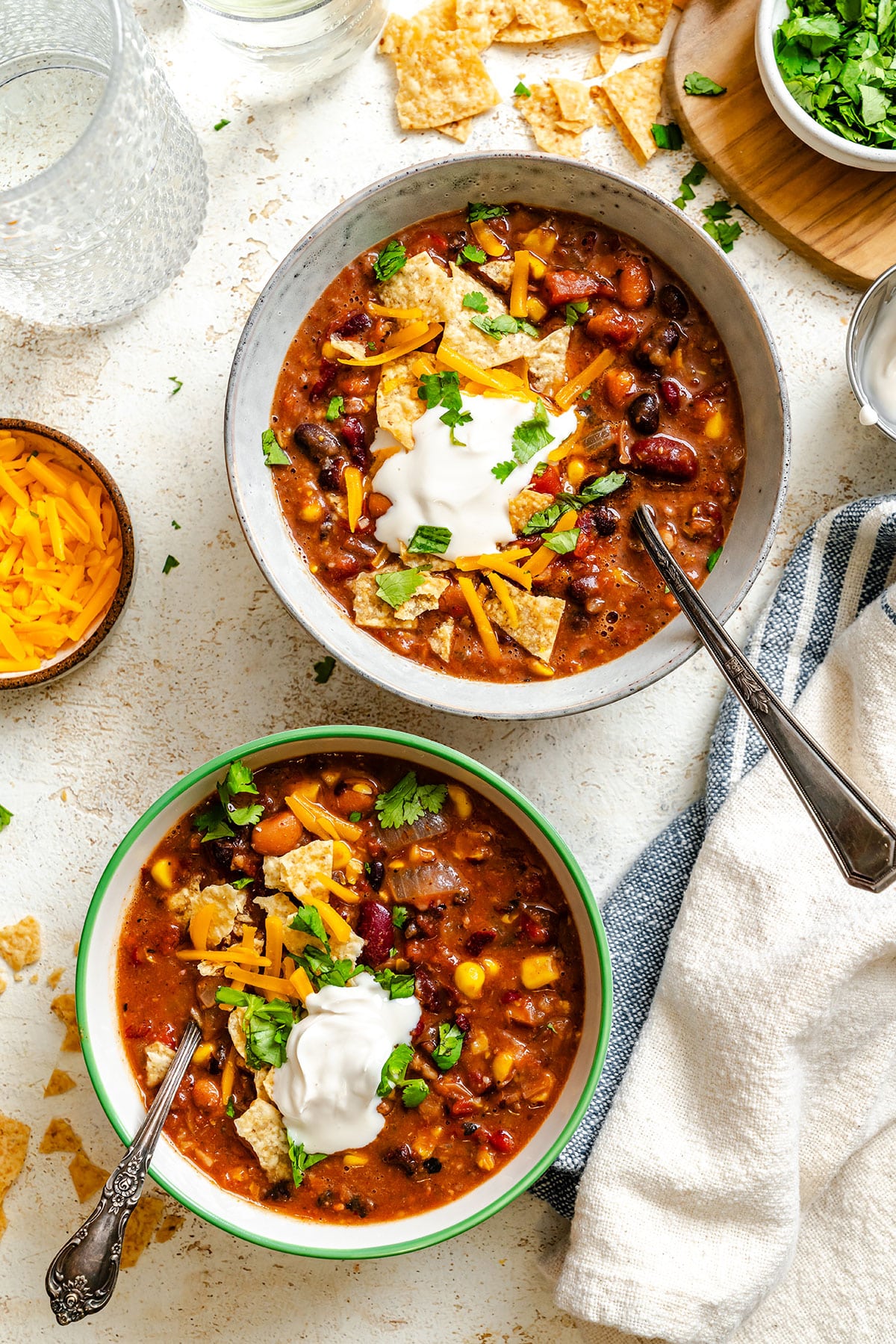 Two bowls of chili topped with sour cream, shredded cheese, tortilla chips, and cilantro sit on a table with a napkin, spoon, and small bowls of toppings nearby.