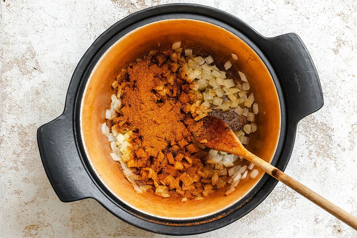 Overhead view of chopped onions and spices in a large pot with a wooden spoon, ready for cooking.