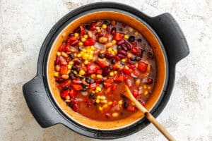 A pot of vegetable chili with beans, corn, tomatoes, and peppers is being stirred with a wooden spoon.