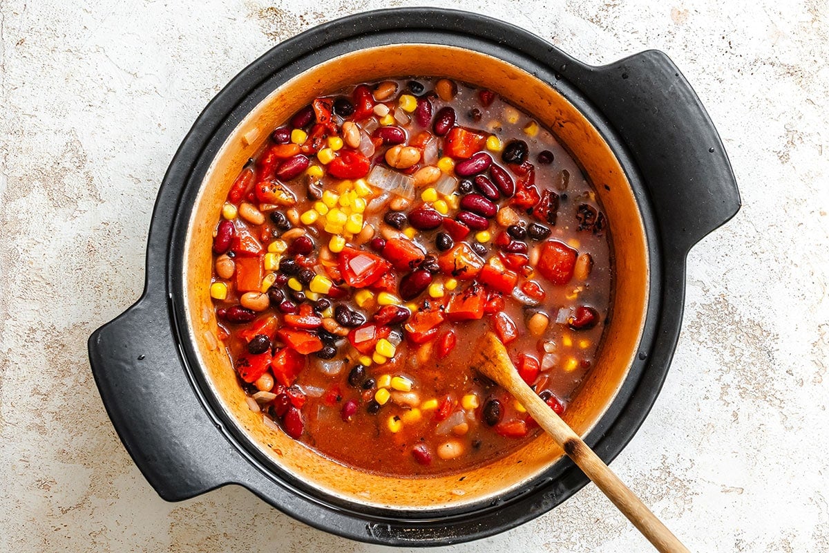 A pot of vegetable chili with beans, corn, tomatoes, and peppers is being stirred with a wooden spoon.