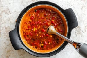 A pot of tomato-based soup with corn and other vegetables is being blended with an immersion blender on a light countertop.