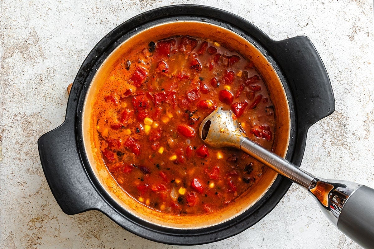 A pot of tomato-based soup with corn and other vegetables is being blended with an immersion blender on a light countertop.