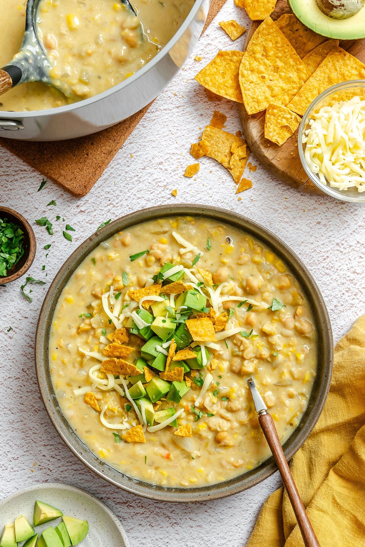 A bowl of creamy soup topped with shredded cheese, chopped avocado, and crushed tortilla chips, with a pot of soup, cheese, chips, and herbs nearby.
