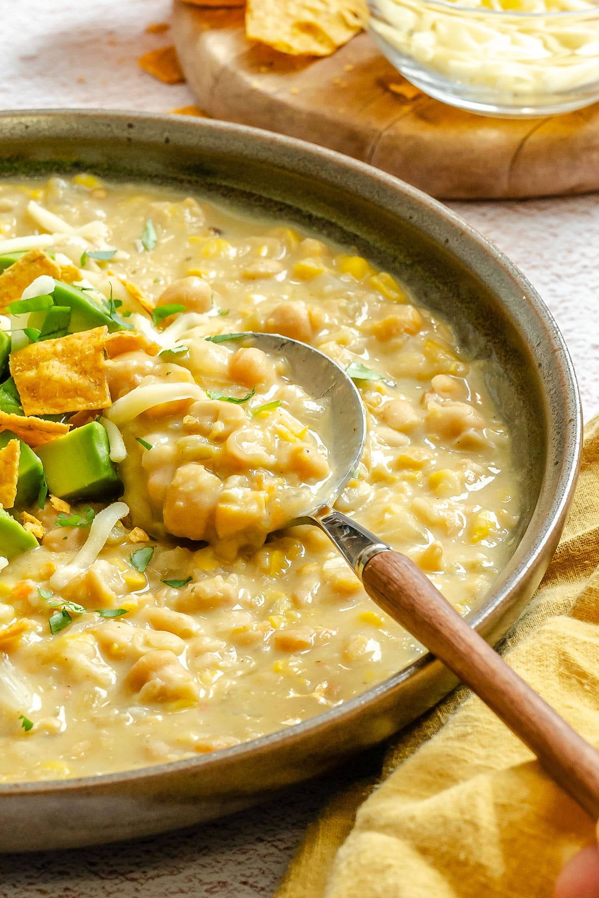 A bowl of creamy white bean chili with corn, topped with avocado, tortilla chips, and shredded cheese, with a spoon in the dish.