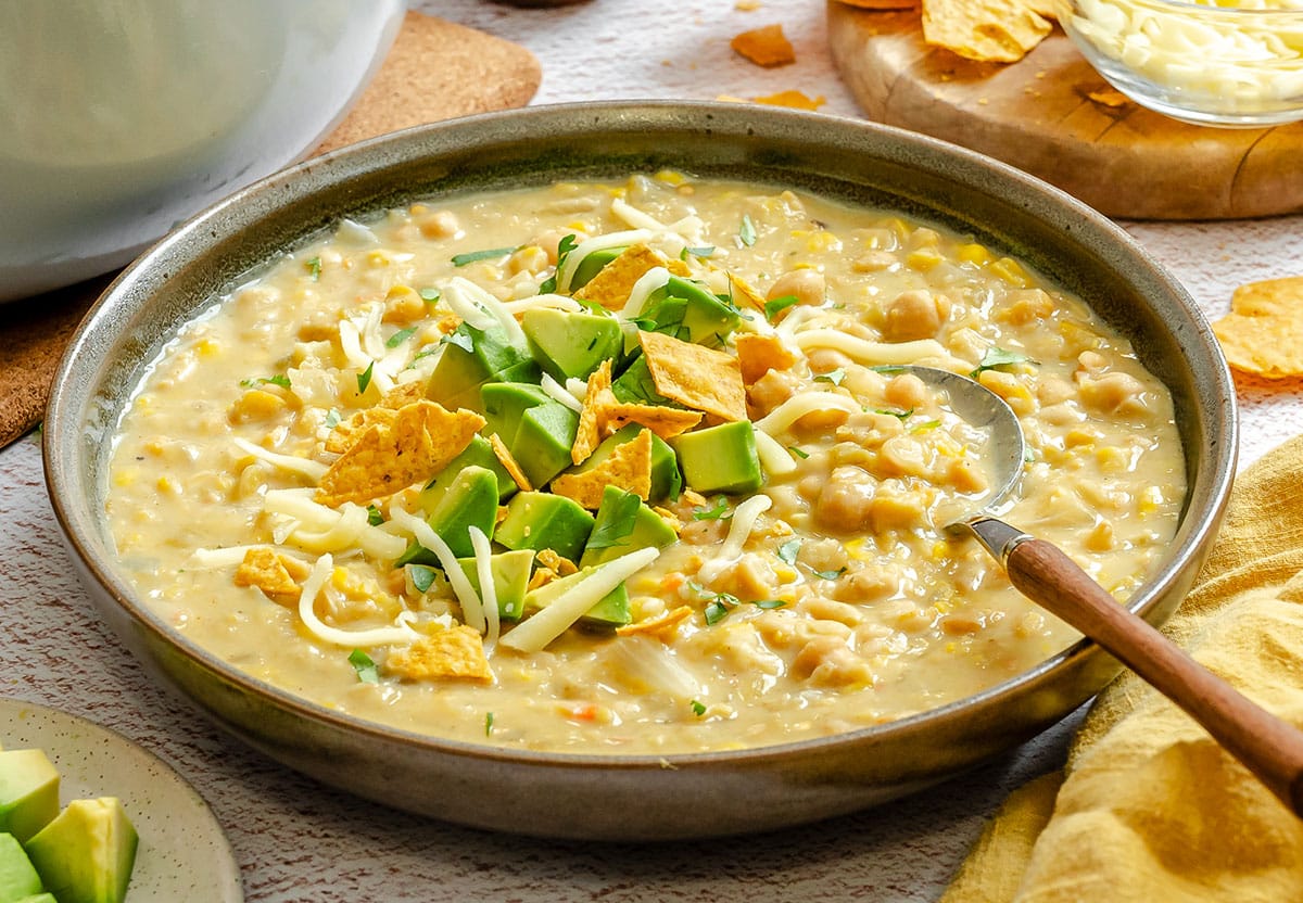 A bowl of creamy white bean chili topped with diced avocado, shredded cheese, and tortilla chips, with a spoon resting in the bowl.