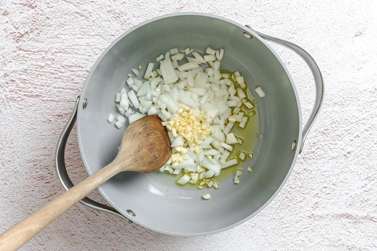 Chopped onions and minced garlic in a gray pot with olive oil, being stirred with a wooden spoon on a light textured surface.