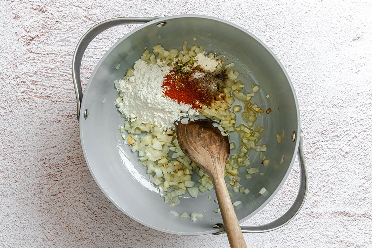 Chopped onions, flour, and assorted spices in a large gray pot with a wooden spoon on a white textured surface.