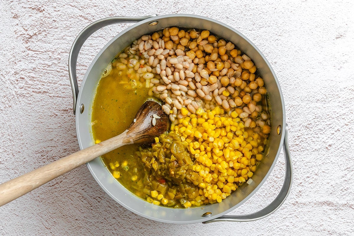 A pot containing white beans, corn, green chilies, broth, and a wooden spoon, viewed from above on a light textured surface.
