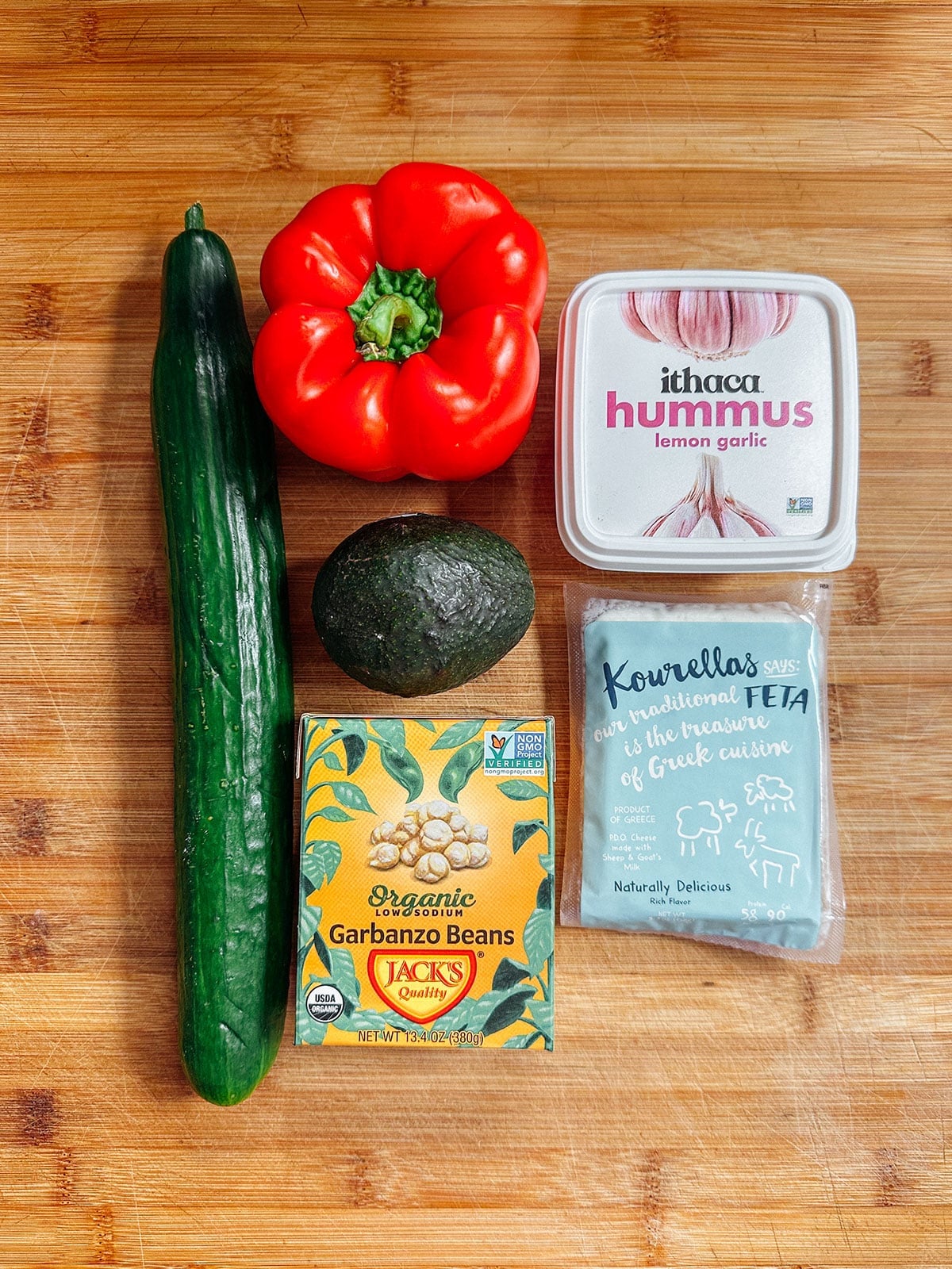 A cucumber, red bell pepper, avocado, box of garbanzo beans, container of lemon garlic hummus, and a package of feta cheese on a wooden surface.