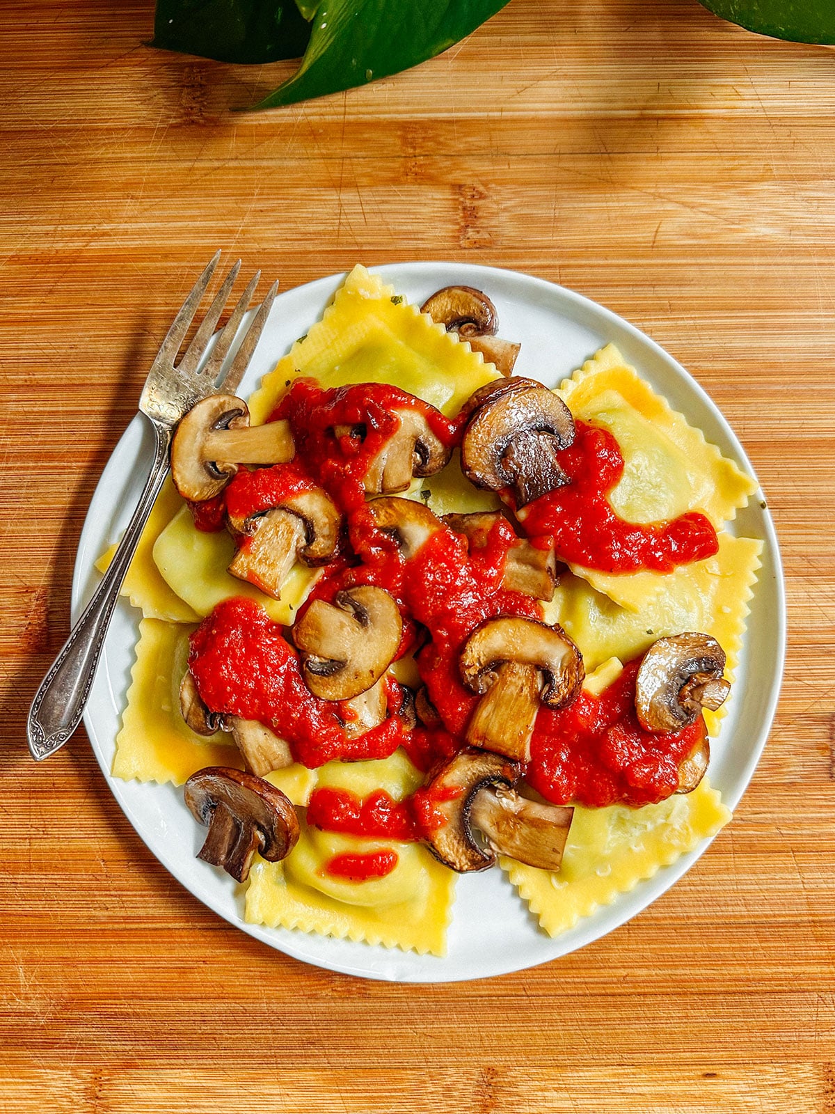 A plate of ravioli topped with tomato sauce and sautéed mushrooms, served with a fork on a wooden surface.