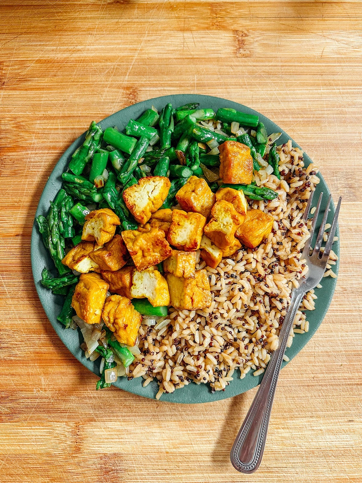 A plate with sautéed green beans, golden tofu cubes, and a mix of brown and wild rice, with a fork on the side, sits on a wooden surface.