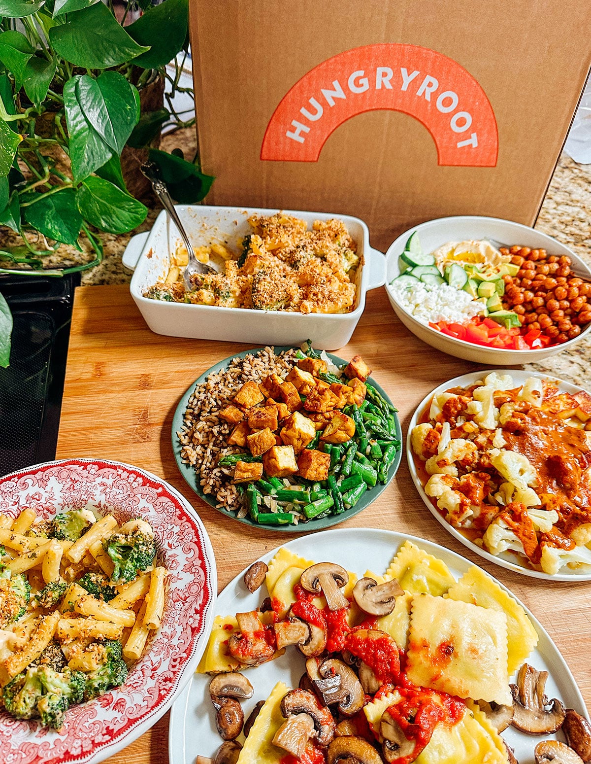 Several colorful plant-based meals are arranged on plates and bowls in front of a large Hungryroot meal delivery box on a wooden surface.