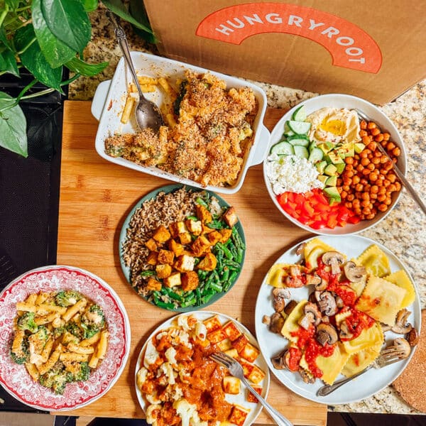 A variety of colorful dishes, including salads, pasta, roasted vegetables, and a casserole, are arranged on a wooden board next to a Hungryroot box on a kitchen counter.