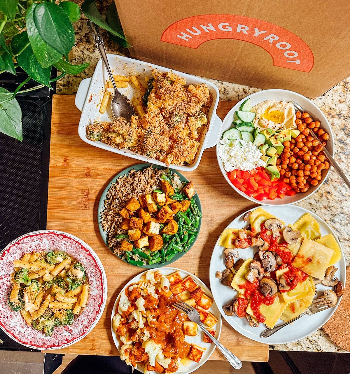 A variety of colorful dishes, including salads, pasta, roasted vegetables, and a casserole, are arranged on a wooden board next to a Hungryroot box on a kitchen counter.