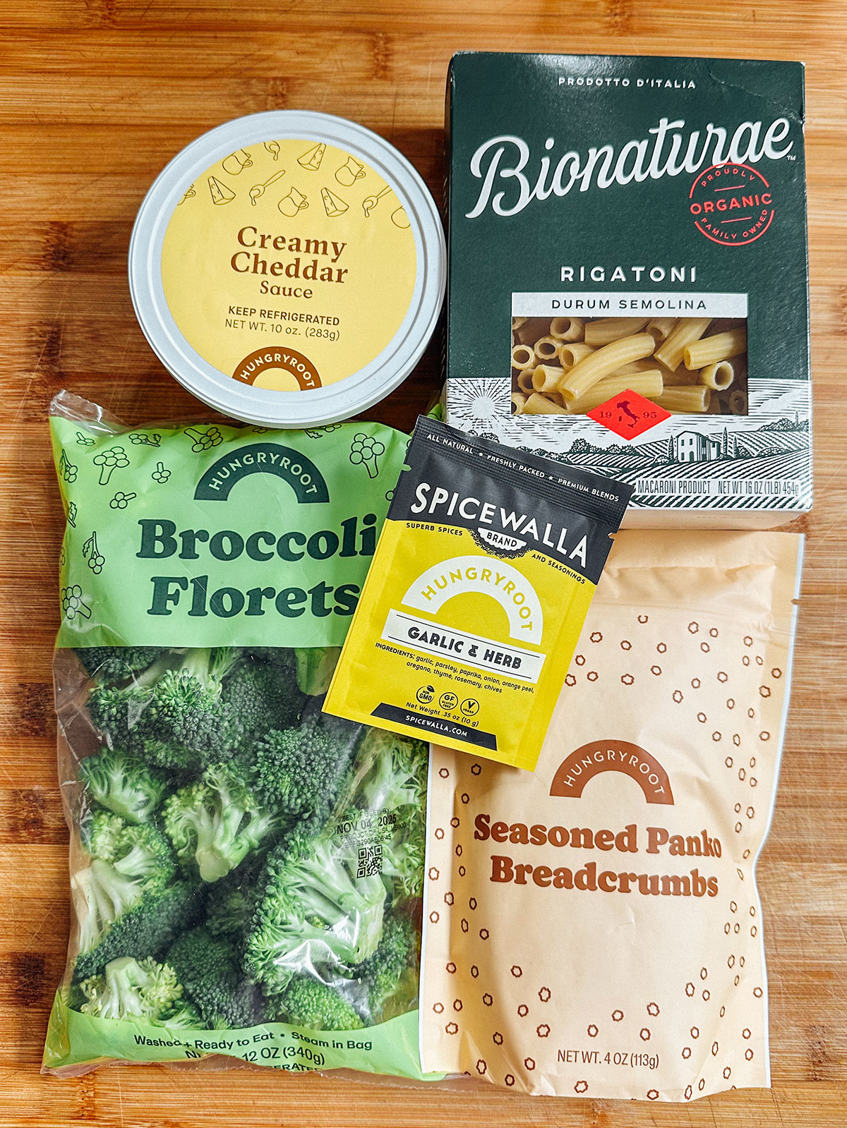 A can of creamy cheddar sauce, a box of rigatoni, a bag of broccoli florets, a packet of garlic herb spice, and a bag of seasoned panko breadcrumbs on a wooden surface.