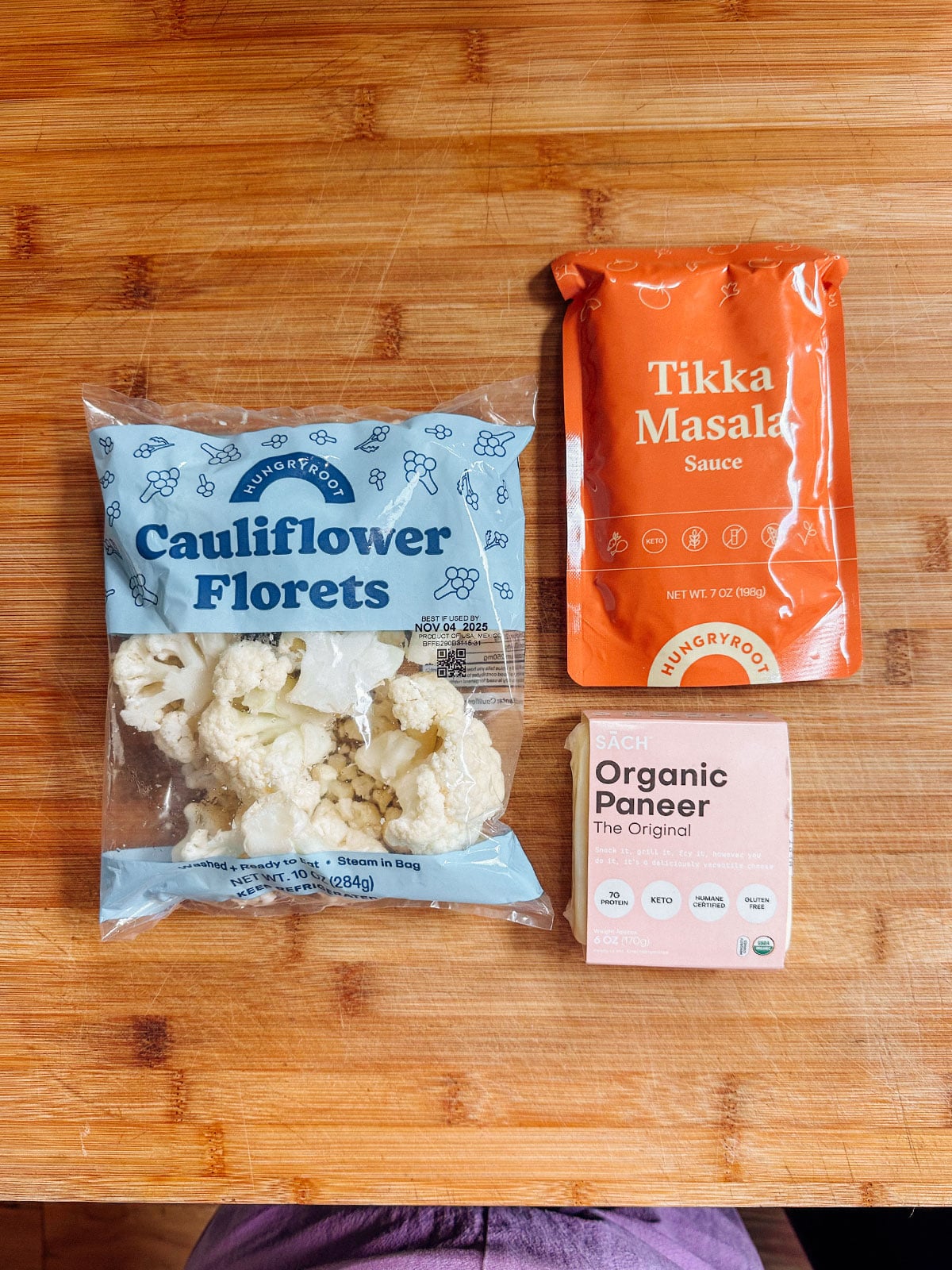 A bag of cauliflower florets, a pack of tikka masala sauce, and a block of organic paneer on a wooden surface.