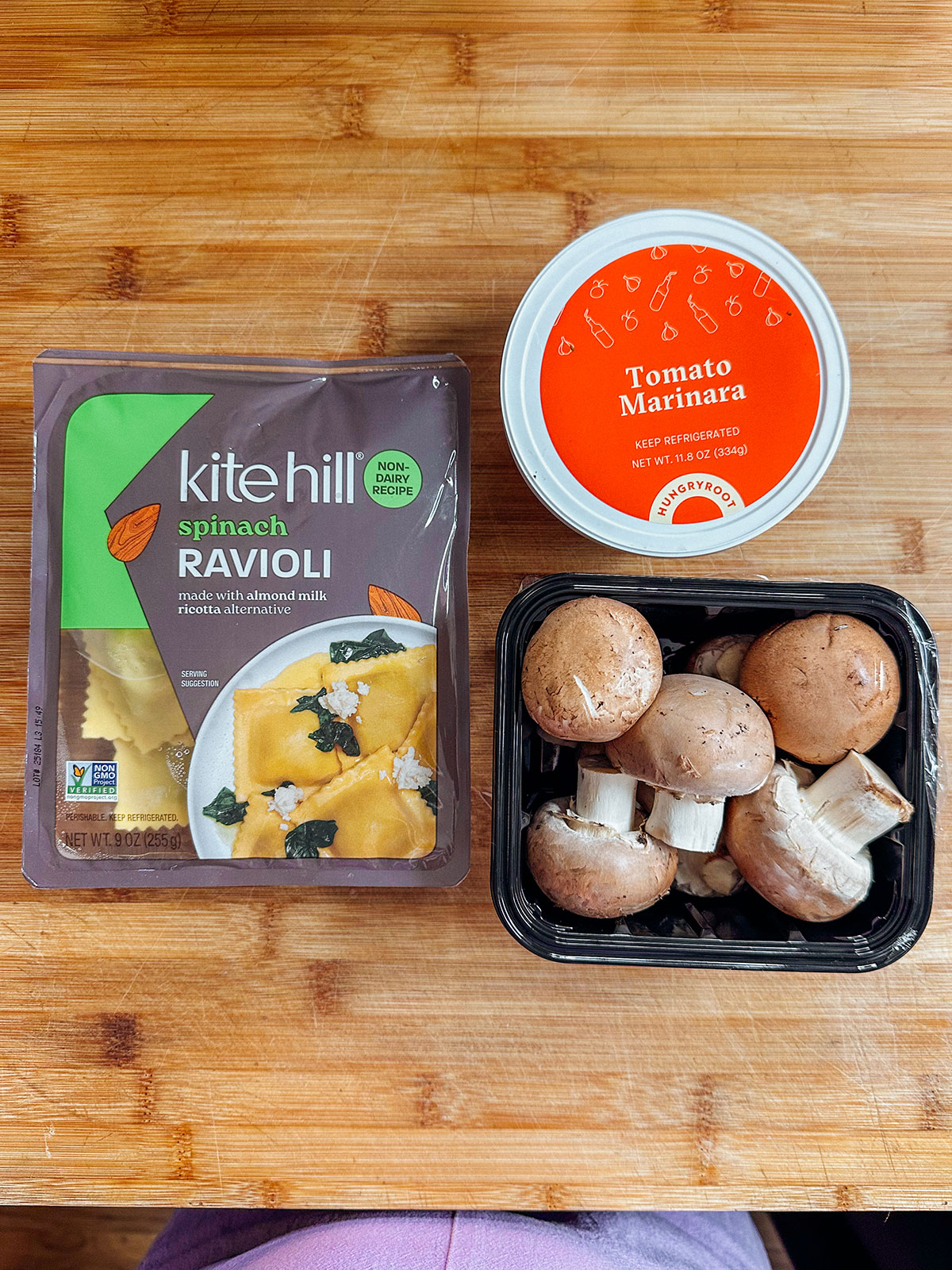 A package of Kite Hill spinach ravioli, a container of tomato marinara sauce, and a black tray of whole cremini mushrooms on a wooden surface.