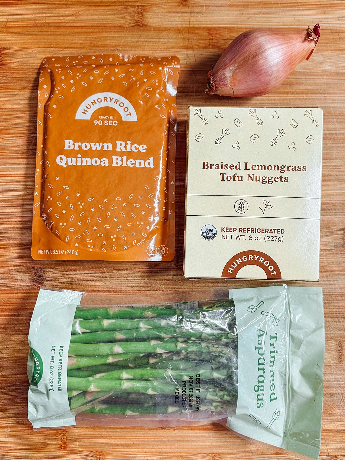 Four food items on a wooden surface: a packet of brown rice quinoa blend, a box of braised lemongrass tofu nuggets, a shallot, and a bag of trimmed asparagus.