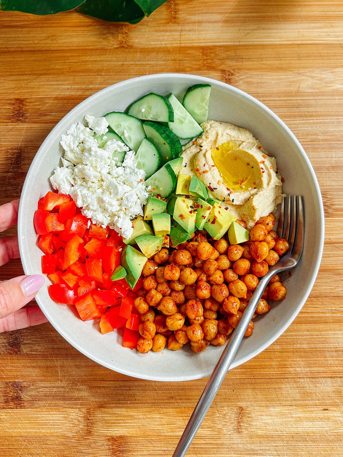 A bowl with sliced cucumber, crumbled feta, diced red bell pepper, avocado, hummus with olive oil, and seasoned chickpeas on a wooden surface, with a fork and a hand holding the bowl.