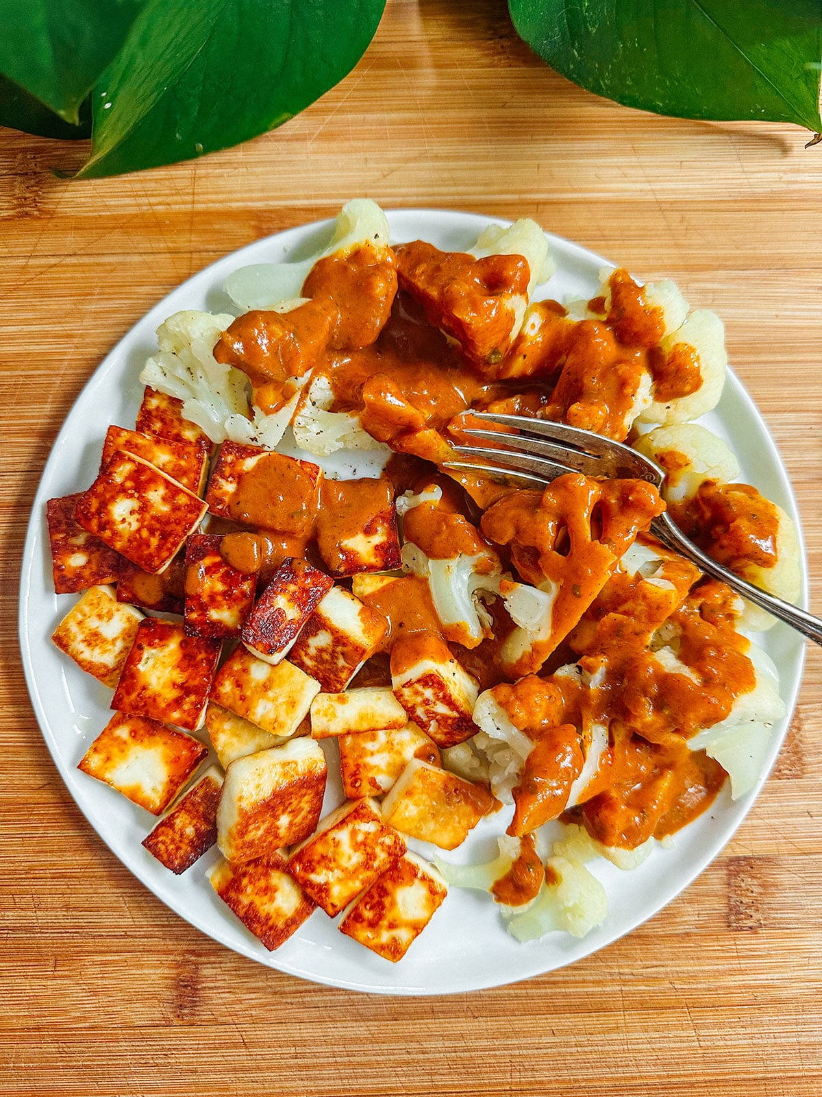 A plate with grilled paneer cubes, steamed cauliflower, and cauliflower topped with brown curry sauce, placed on a wooden surface. A fork rests on the plate.