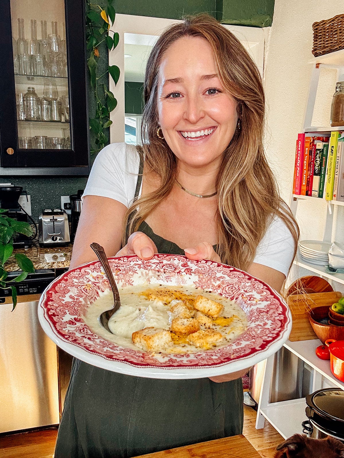 A woman stands in a kitchen, smiling and holding a bowl of creamy soup with croutons and a spoon, presented on a decorative red and white plate.